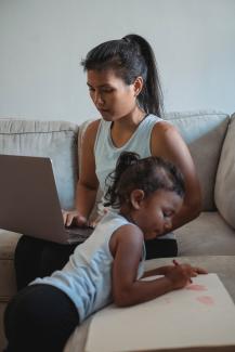 A mom works on her laptop with her young child coloring nearby.