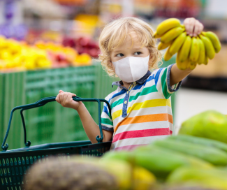 A young boy with blond hair masked and shopping for groceries with family. He is holding up bananas. 
