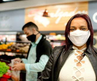 A close up of a woman, with a mask, in an off white shirt and leather jacket is food shopping at a grocery store. 