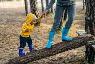 In colorful rain boots, a parent helps a toddler up a ramp made of a wooden log that is in a park.