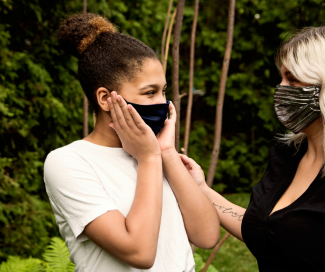 A woman touches the shoulder of another woman to console her as they stand in a park. Both are masked. 
