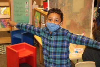 A boy with a mask and a checkered shirt stretches out his arms wide while in child care. 