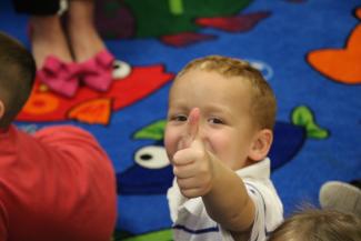 A little boy sitting on a colorful run at a child care site gives a thumbs up.