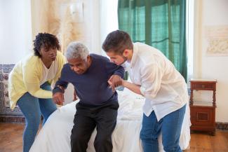 A young man and woman help an older man with standing up from a bed. 