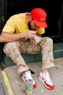 A man with camouflage pants, yellow shirt and a red baseball cap sits to take a quick bite of food. His drink is on the ground next to his sneakers.