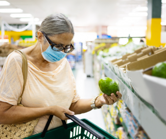 An older woman with a mask adds a green pepper to her grocery shopping basket. 