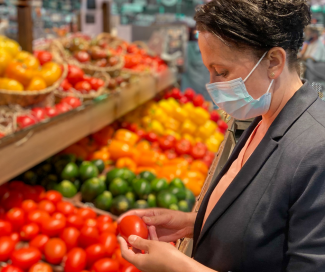 A woman with a mask and her hair pinned is selecting a tomato in the produce section of a grocery store. 