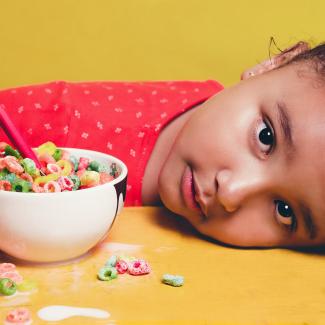 Staring directly at the camera, a school age child has her head on a kitchen table. Next to her is a bowl of fruity cereal, including some loops and milk that spilled onto the table. 