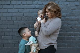 A mom holding her infant child looks down at her son who is looking back up at her. They are standing in front of a grey brick building. 