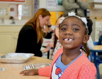 A little girl with a pick sweater, Hello Kitty shirt and missing front teeth smiles while sitting at a table in a child care site. 