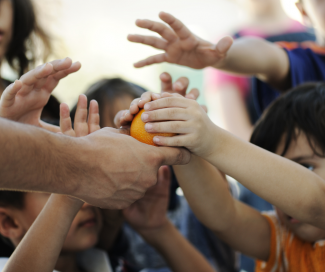 The hands of a few children reaching for an orange.