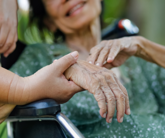 A person holds the hand of an older woman who is smiling and seated in a wheelchair.