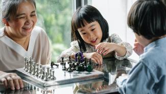A grandmother happily plays chess with her visiting grandchildren.
